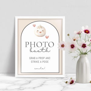 May include: A framed photo booth sign with a cartoon dumpling illustration and the words "PHOTO booth." The sign also includes the phrases "Grab a prop and strike a pose" and "smile!" A vase of white and pink flowers is to the right.