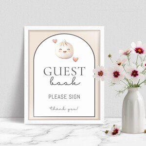 May include: A framed guest book sign with a white border. The sign features a smiling dumpling illustration and the words "GUEST book PLEASE SIGN thank you!" A vase of white and pink flowers sits to the right.