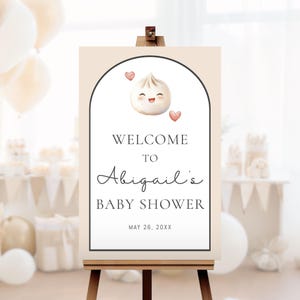 May include: A baby shower welcome sign on an easel. The sign has a beige background with a white arch containing a smiling dumpling and two hearts. Text reads "Welcome to Abigail's Baby Shower" and the date.