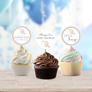 May include: Three cupcakes with different frosting colors and a round, white, paper cupcake topper with a cartoon dumpling and the text "A little dumpling is on the way!" on one, a round, white, paper cupcake topper with a cartoon dumpling and the text "Abigail's Baby Shower" on another, and a round, white, paper cupcake topper with a cartoon dumpling and the text "It's a boy" on the third.