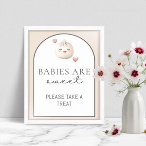 May include: A framed sign with the text "BABIES ARE sweet PLEASE TAKE A TREAT". The sign features a cartoon dumpling with a smiling face and two pink hearts. The sign is in a white frame, with a vase of flowers to the right.