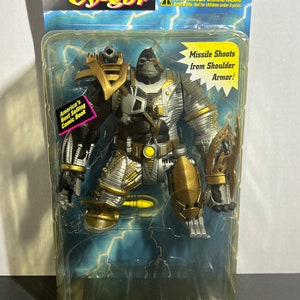 May include: A Cy-Gor action figure in a plastic package. The figure is a gorilla wearing silver and gold armor. The figure has a missile launcher on its shoulder. The package has a warning label that says "WARNING: CHOKING HAZARD. Small Parts. Not for children under 3 years."