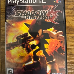 Puede incluir: Una caja de videojuego de PlayStation 2 para Shadow the Hedgehog que muestra al personaje negro sosteniendo un arma de fuego sobre un fondo naranja y negro ardiente. La portada incluye el título del juego, el logo de Sega y la clasificación ESRB Everyone 10+.