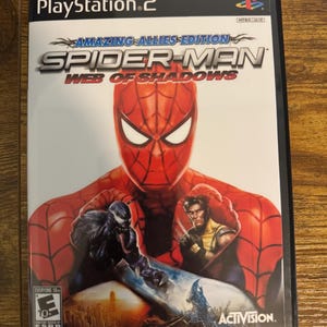 Puede incluir: Una caja de videojuego de PlayStation 2 para Spider-Man: Web of Shadows Amazing Allies Edition. La portada muestra una gran máscara roja de Spider-Man, Venom y Wolverine. La clasificación ESRB es Everyone 10+ y dice Reprinted Art.
