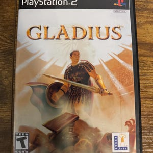 Peut inclure: Un boîtier de jeu vidéo PlayStation 2 pour le titre Gladius. La jaquette représente un guerrier tenant une épée et un bouclier, avec le texte Gladius, PlayStation 2, LucasArts et une classification ESRB Teen. Le bas indique Reprinted Art.
