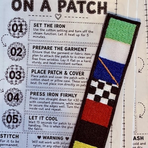 May include: A rectangular embroidered patch featuring stacked horizontal color blocks of green, white, blue with an orange diagonal line, black and white checkerboard, red, black, and yellow, placed on an instruction sheet titled HOW TO IRON ON A PATCH.