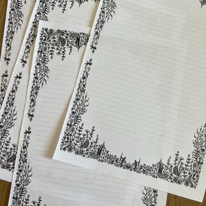 Peut inclure: Trois feuilles de papier &agrave; lettres blanc avec des bordures botaniques dessin&eacute;es &agrave; l'encre noire. Chaque page pr&eacute;sente des lignes horizontales pour l'&eacute;criture, entour&eacute;es d'illustrations complexes de vignes, de feuilles et de fleurs.