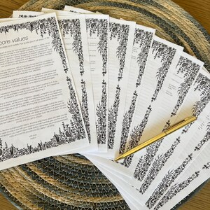 May include: A stack of white paper sheets with black printed text titled core values and intricate floral border designs, accompanied by a gold-colored ballpoint pen. The pages feature lined sections for writing and decorative botanical illustrations.