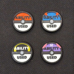 May include: Four round, black and white tokens with a Pokeball design. Each token has a different colored rim: orange, blue, yellow, and purple. The text "ABILITY USED" is printed on each token.