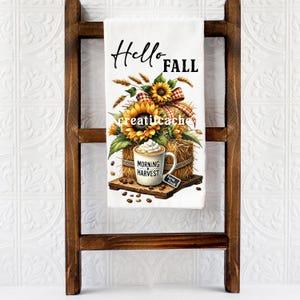 May include: White kitchen towel with a fall-themed design. The towel features the words "Hello Fall" and a graphic of sunflowers, a coffee mug with whipped cream, and a hay bale. The towel is displayed on a wooden ladder.