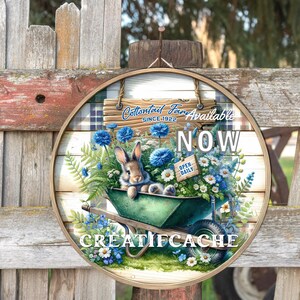 May include: A round wooden sign with a white background and a blue and white checkered pattern. The sign says "Cottontail Farm Available Now" with a bunny rabbit sitting in a green wheelbarrow filled with blue and white flowers. The sign also says "Open Daily" and "Creatifcache".