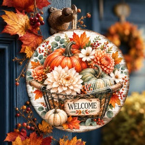May include: A round wooden sign featuring a wire basket filled with orange and green pumpkins, white and orange dahlias, and autumn maple leaves. A rectangular wooden plaque attached to the front of the basket displays the word WELCOME in black text.