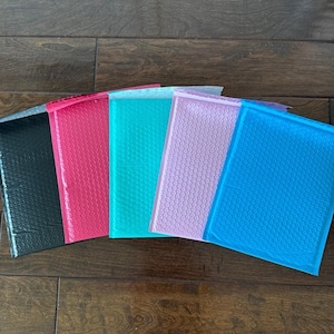 Puede incluir: Una colección de sobres de burbujas de colores en varios tonos. Los sobres incluyen negro, rosa intenso, verde azulado, rosa claro y azul. Cada sobre tiene una superficie texturizada de plástico de burbujas, diseñada para amortiguar y proteger los artículos durante el envío.