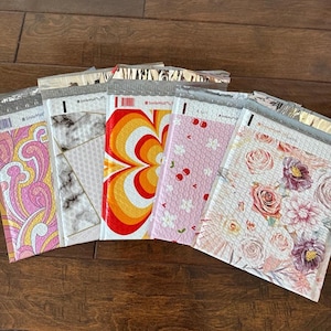 Puede incluir: Una colección de sobres de burbujas coloridos con varios diseños. Los patrones incluyen un diseño psicodélico rosa y amarillo, mármol, un remolino retro naranja y amarillo, cerezas rosas e impresiones florales. Los sobres están sellados con una tira plateada.
