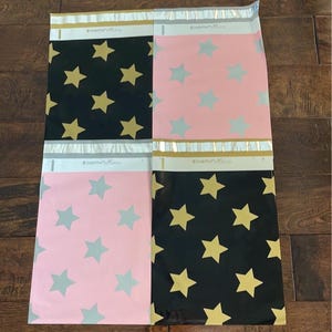 May include: Four colorful poly mailer envelopes. Two are black with gold stars, and two are pink with gray stars. The envelopes are sealed at the top with a silver and gold strip. The envelopes are on a wooden surface.
