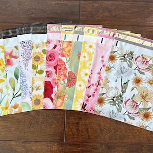 Può includere: Una collezione di buste per la spedizione in polietilene con motivi floreali. I disegni includono girasoli, rose, margherite e altri fiori. Le buste sono disposte su una superficie di legno scuro.