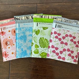 May include: A collection of colorful bubble mailers with various patterns. Designs include pink daisies, blue tie-dye, green tropical leaves, and red cherries. Each mailer has a metallic silver top and the text "#SmileMail by UpInShop".