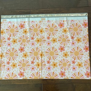 May include: White poly mailers with a repeating pattern of pink flowers and orange sunbursts. The mailers have a silver stripe across the top.