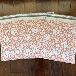 Könnte beinhalten: Mehrere Papierbögen mit einem sich wiederholenden Muster aus weißen Pfotenabdrücken auf korallenfarbenem Hintergrund. Die Bögen sind in klarem Kunststoff verpackt. Das Bild befindet sich auf einer dunklen Holzoberfläche.