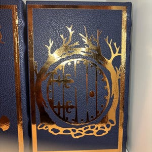 K&ouml;nnte beinhalten: Marineblaues Buch mit einem goldfarbenen Design aus Tor und B&auml;umen. Der Einband hat eine strukturierte Optik und einen goldenen Rahmen. Das Buch ist rechteckig, m&ouml;glicherweise ein Tagebuch oder Notizbuch.