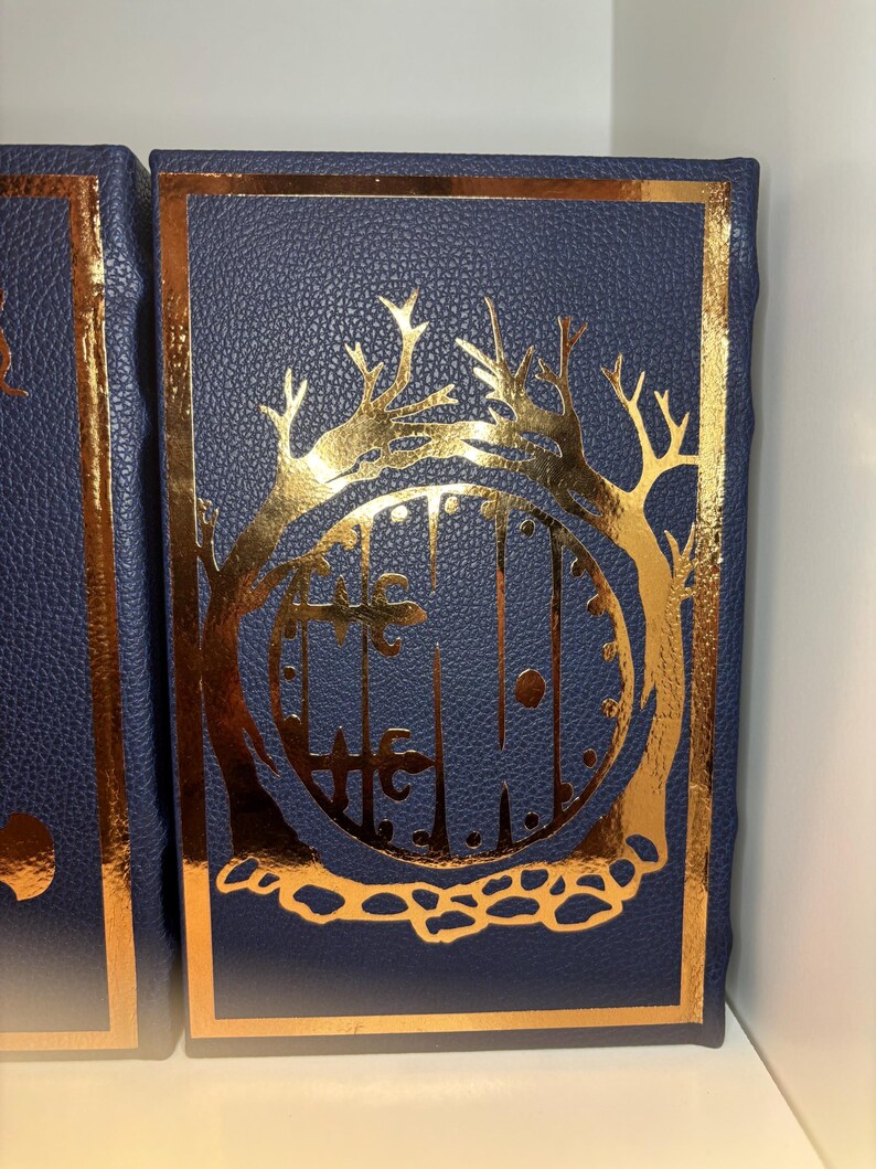 K&ouml;nnte beinhalten: Marineblaues Buch mit einem goldfarbenen Design aus Tor und B&auml;umen. Der Einband hat eine strukturierte Optik und einen goldenen Rahmen. Das Buch ist rechteckig, m&ouml;glicherweise ein Tagebuch oder Notizbuch.