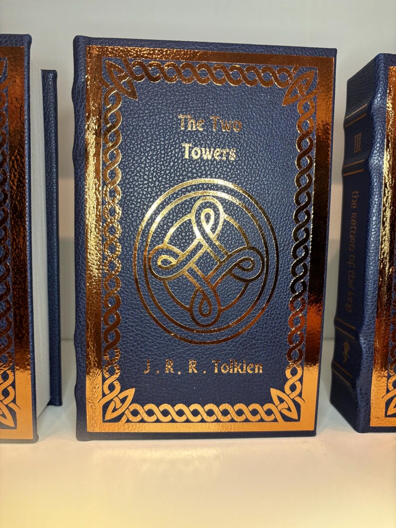 K&ouml;nnte beinhalten: Ein marineblaues Buch mit dem Titel "The Two Towers" von J.R.R. Tolkien, mit goldenen Details. Das Cover zeigt ein goldenes Muster und einen Rand. Der Buchr&uuml;cken ist ebenfalls sichtbar, mit dem Titel "The Return of the King".