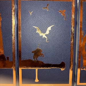 K&ouml;nnte beinhalten: Ein Satz von drei blau ledergebundenen B&uuml;chern mit goldenen Folienakzenten. Die Einb&auml;nde zeigen einen Drachen, V&ouml;gel und einen Ritter auf einer Kanone. Die Goldfolie bildet einen auff&auml;lligen Kontrast zum dunkelblauen Hintergrund.