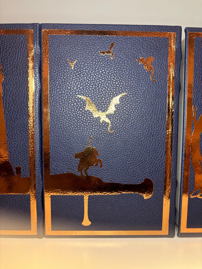 K&ouml;nnte beinhalten: Ein Satz von drei blau ledergebundenen B&uuml;chern mit goldenen Folienakzenten. Die Einb&auml;nde zeigen einen Drachen, V&ouml;gel und einen Ritter auf einer Kanone. Die Goldfolie bildet einen auff&auml;lligen Kontrast zum dunkelblauen Hintergrund.