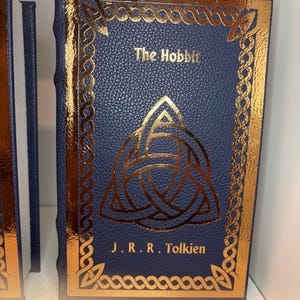 K&ouml;nnte beinhalten: Ein gebundenes Buch mit dem Titel "The Hobbit" von J.R.R. Tolkien. Das Buch hat einen dunkelblauen Einband mit goldfarbenen Akzenten, darunter ein keltisches Knotenmuster und dekorative Bord&uuml;ren. Der Buchr&uuml;cken ist ebenfalls dunkelblau.