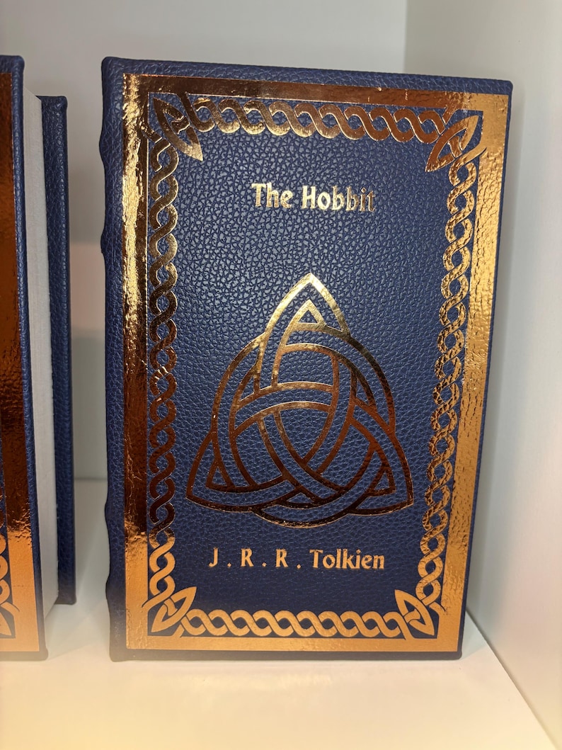 K&ouml;nnte beinhalten: Ein gebundenes Buch mit dem Titel "The Hobbit" von J.R.R. Tolkien. Das Buch hat einen dunkelblauen Einband mit goldfarbenen Akzenten, darunter ein keltisches Knotenmuster und dekorative Bord&uuml;ren. Der Buchr&uuml;cken ist ebenfalls dunkelblau.