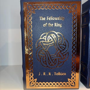 K&ouml;nnte beinhalten: Ein blaues Hardcover-Buch mit dem Titel "The Fellowship of the Ring" von J.R.R. Tolkien. Das Cover zeigt goldfarbene keltische Knotenmuster und Text. Das Buch hat einen goldenen Rand und ein blaues Lesezeichen.