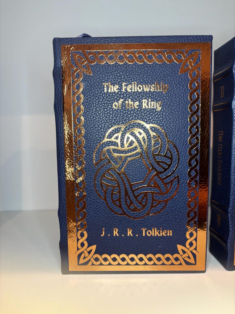 K&ouml;nnte beinhalten: Ein blaues Hardcover-Buch mit dem Titel "The Fellowship of the Ring" von J.R.R. Tolkien. Das Cover zeigt goldfarbene keltische Knotenmuster und Text. Das Buch hat einen goldenen Rand und ein blaues Lesezeichen.