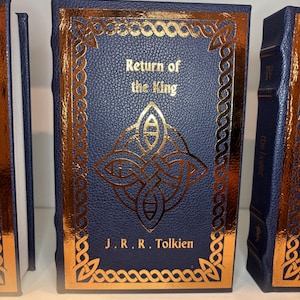 K&ouml;nnte beinhalten: Eine Sammlung von Hardcover-B&uuml;chern mit marineblauen Einb&auml;nden und goldenen Folienakzenten. Das zentrale Buch tr&auml;gt den Titel "Return of the King" von J.R.R. Tolkien und zeigt ein goldenes keltisches Knotenmuster. Die Buchkanten sind ebenfalls vergoldet.