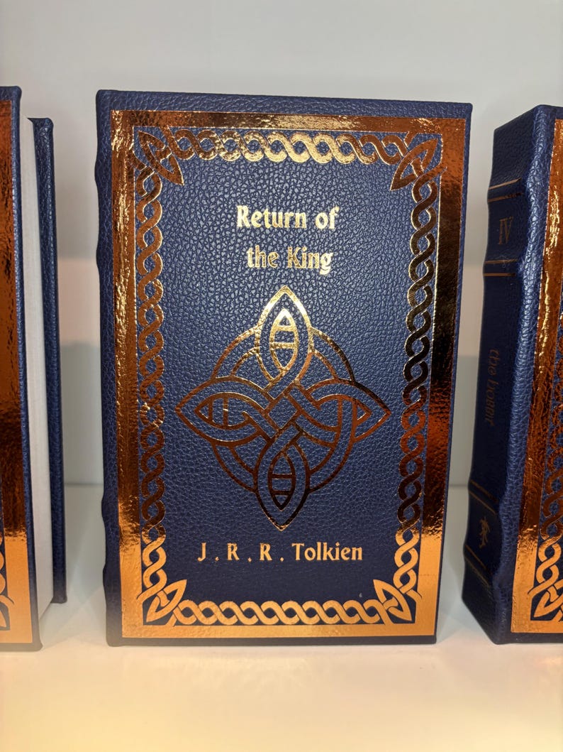 K&ouml;nnte beinhalten: Eine Sammlung von Hardcover-B&uuml;chern mit marineblauen Einb&auml;nden und goldenen Folienakzenten. Das zentrale Buch tr&auml;gt den Titel "Return of the King" von J.R.R. Tolkien und zeigt ein goldenes keltisches Knotenmuster. Die Buchkanten sind ebenfalls vergoldet.