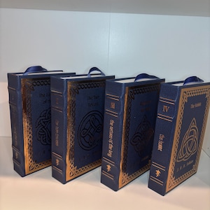 K&ouml;nnte beinhalten: Vier blaue Hardcover-B&uuml;cher mit goldenen Verzierungen und keltischen Knotenmustern. Die B&uuml;cher haben blaue Leseb&auml;ndchen. Titel sind "The Fellowship of the Ring", "The Two Towers", "The Return of the King" und "The Hobbit".
