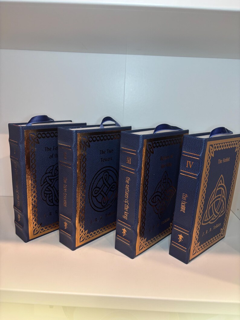 K&ouml;nnte beinhalten: Vier blaue Hardcover-B&uuml;cher mit goldenen Verzierungen und keltischen Knotenmustern. Die B&uuml;cher haben blaue Leseb&auml;ndchen. Titel sind "The Fellowship of the Ring", "The Two Towers", "The Return of the King" und "The Hobbit".