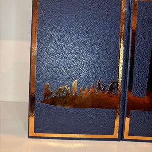 K&ouml;nnte beinhalten: Zwei marineblaue Boxen mit Lederstruktur, goldfarbenem Rand und goldenem Silhouette-Design. Die Silhouette zeigt eine Landschaft mit Figuren. Die Boxen stehen nebeneinander, die rechte Box ist teilweise sichtbar.