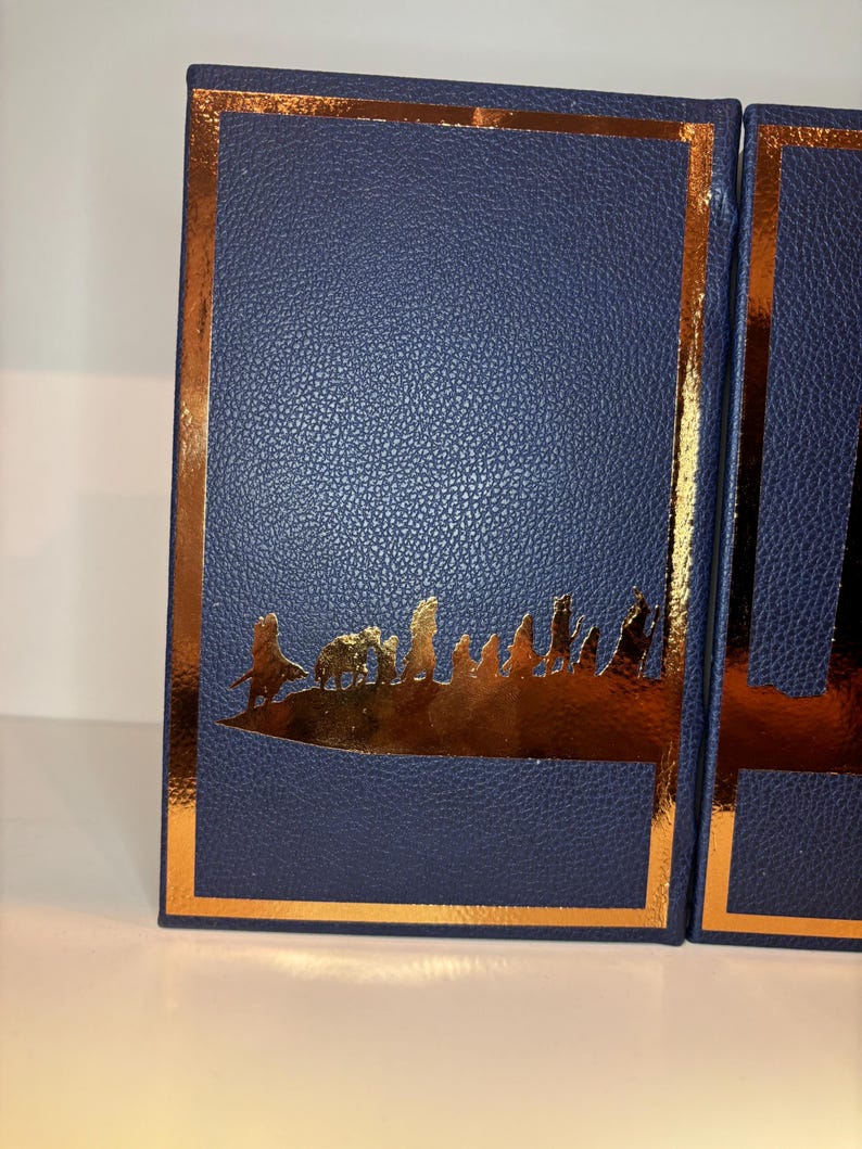 K&ouml;nnte beinhalten: Zwei marineblaue Boxen mit Lederstruktur, goldfarbenem Rand und goldenem Silhouette-Design. Die Silhouette zeigt eine Landschaft mit Figuren. Die Boxen stehen nebeneinander, die rechte Box ist teilweise sichtbar.