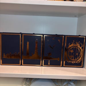 K&ouml;nnte beinhalten: Vier marineblaue Buchumschl&auml;ge mit goldfarbenen Folienaussparungen von Landschaften und architektonischen Designs. Die Umschl&auml;ge sind nebeneinander auf einem wei&szlig;en Regal angeordnet. Die Designs umfassen eine Stadtsilhouette, einen Leuchtturm, ein fliegendes Wesen und eine Hobbit-T&uuml;r.