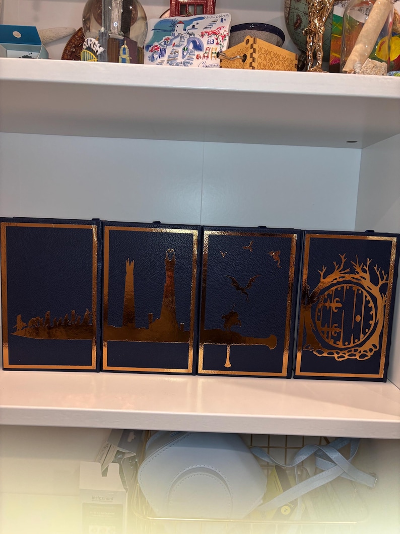 K&ouml;nnte beinhalten: Vier marineblaue Buchumschl&auml;ge mit goldfarbenen Folienaussparungen von Landschaften und architektonischen Designs. Die Umschl&auml;ge sind nebeneinander auf einem wei&szlig;en Regal angeordnet. Die Designs umfassen eine Stadtsilhouette, einen Leuchtturm, ein fliegendes Wesen und eine Hobbit-T&uuml;r.