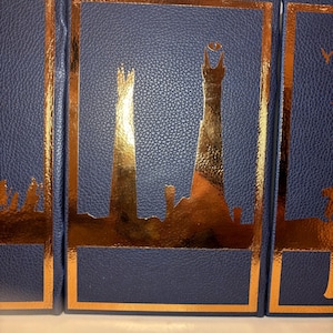 K&ouml;nnte beinhalten: Drei marineblaue B&uuml;cher mit goldfarbenen Details. Jedes Buch hat ein goldfarbenes Skyline-Design innerhalb eines goldenen Rahmens. Die B&uuml;cher haben eine strukturierte, lederartige Optik.