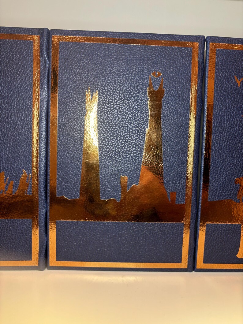 K&ouml;nnte beinhalten: Drei marineblaue B&uuml;cher mit goldfarbenen Details. Jedes Buch hat ein goldfarbenes Skyline-Design innerhalb eines goldenen Rahmens. Die B&uuml;cher haben eine strukturierte, lederartige Optik.