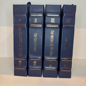 K&ouml;nnte beinhalten: Vier marineblaue Hardcover-B&uuml;cher mit goldfarbenen Buchstaben und Verzierungen. Die Buchr&uuml;cken zeigen r&ouml;mische Ziffern I bis IV und Titel. Die B&uuml;cher sind nebeneinander angeordnet, was auf ein Set hindeutet.