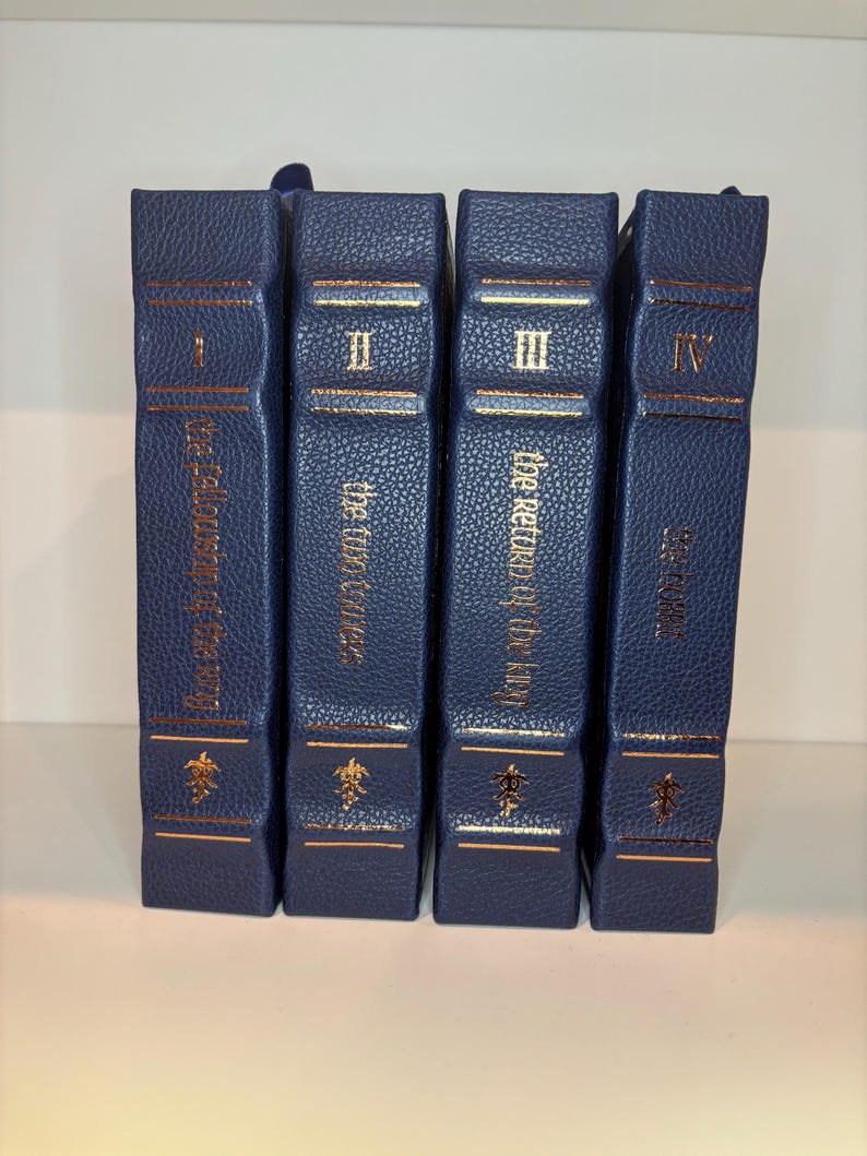 K&ouml;nnte beinhalten: Vier marineblaue Hardcover-B&uuml;cher mit goldfarbenen Buchstaben und Verzierungen. Die Buchr&uuml;cken zeigen r&ouml;mische Ziffern I bis IV und Titel. Die B&uuml;cher sind nebeneinander angeordnet, was auf ein Set hindeutet.