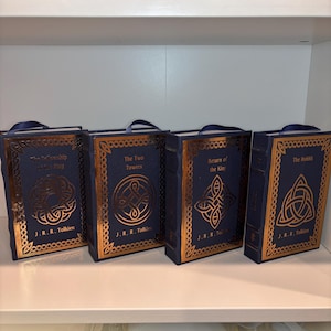 K&ouml;nnte beinhalten: Vier blaue, buchf&ouml;rmige Schachteln mit goldgepr&auml;gten Designs und Titeln aus J.R.R. Tolkiens Werken: "Die Gef&auml;hrten", "Die zwei T&uuml;rme", "Die R&uuml;ckkehr des K&ouml;nigs" und "Der Hobbit". Jede hat einen blauen Bandgriff.
