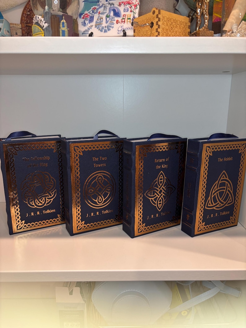 K&ouml;nnte beinhalten: Vier blaue, buchf&ouml;rmige Schachteln mit goldgepr&auml;gten Designs und Titeln aus J.R.R. Tolkiens Werken: "Die Gef&auml;hrten", "Die zwei T&uuml;rme", "Die R&uuml;ckkehr des K&ouml;nigs" und "Der Hobbit". Jede hat einen blauen Bandgriff.