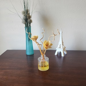 May include: A clear glass diffuser bottle with a yellow liquid and wooden sticks. The bottle is on a wooden table with a blue vase and a white Eiffel Tower figurine.