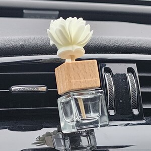 Peut inclure: Un diffuseur d'air pour voiture avec un dessus en forme de fleur blanche, un bloc de bois et une bouteille en verre transparente. Le diffuseur est monté sur une grille d'aération de voiture. L'intérieur de la voiture est noir, et le logo Jaguar est visible.