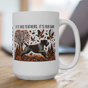 May include: White ceramic mug featuring a hunting dog illustration with the text "IF IT HAS FEATHERS, IT'S FAIR GAME." The design includes a brown and white dog, birds, and autumn foliage. The mug has a large handle and a glossy finish.
