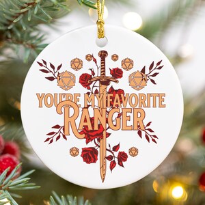 May include: White ceramic Christmas ornament with a gold string, featuring the text "YOU'RE MY FAVORITE RANGER" in orange, with a sword and floral design. The ornament is decorated with red roses and geometric shapes.